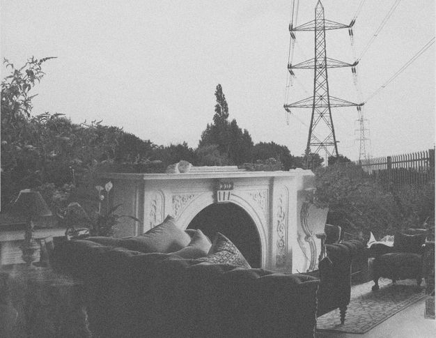 Black and white image. A couch, fireplace mantle, chairs, and an area rug are juxtaposed, perhaps collaged, against a landscape of trees and power lines.