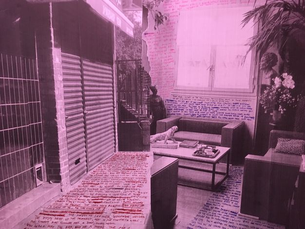 Installation. What initially looks like a single black and white photograph tinted magenta is two views, one of a shuttered storefront and one of the inside of a room with couches and a coffee table. Text in red, blue, or pink appears to have been written on the pavement in front of the store and the floor and walls of the room.