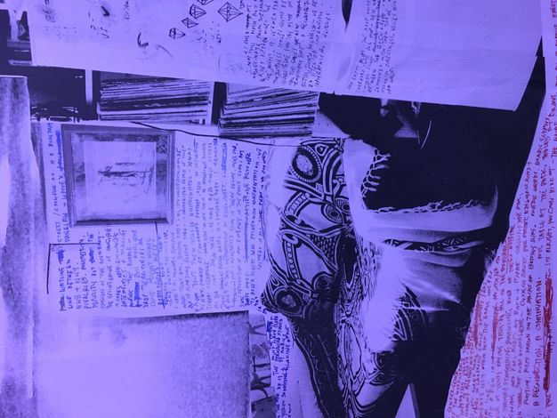 Installation. A black and white photograph tinted delphinium blue is of a room with a blanket-covered couch. Red or blue text has been written on the walls and floor.