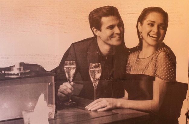 Installation. A peach-tinted black and white image shows a man and woman smiling and holding glasses of champagne. They have been removed from their original context and shown against a background with tiny red writing.