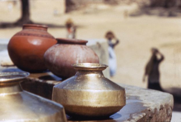 Two people carry vessels balanced on head pads. Both wear scarves covering their heads, and carry red-coloured pots. The person to our left, who faces us, carries a gold vessel balanced on top of the red one.