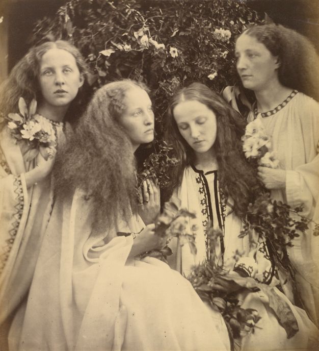 Black and white photograph. Four pale-skinned women are posed closely together in a garden. All have long hair parted down the middle and brushed into puffy clouds. Looking in all directions, they wear long, light-coloured robes and hold flowers.