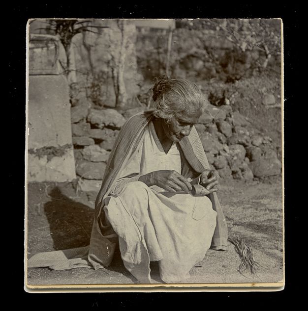 Black and white photograph. Eight people stand on either side of long threads that stretch across at least three thatched houses at waist height. One person bends over the threads. All wear cloth wrapped around their hips or draped over their shoulders. A pencil inscription below the image reads, “Plate I, Weavers”.