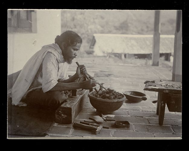 Black and white photograph. A dark-skinned woman with greying hair pulled back from her wrinkled forehead sits low to or on the ground in front of a stone wall. She works on an object, perhaps weaving, balanced on her knees.